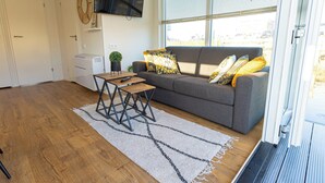 A modern living room with a grey sofa, a wooden coffee table, and a large window with blinds. - Houseboat With Rooftop Terrace (Wolphaartsdijk)