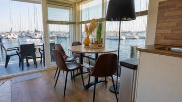 Un comedor con una mesa y sillas, un jarrón con plantas secas y vistas a un puerto deportivo con barcos a través de amplios ventanales.