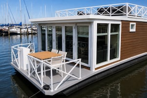 Una casa flotante con zona de comedor, barandillas blancas y puertas de vidrio.