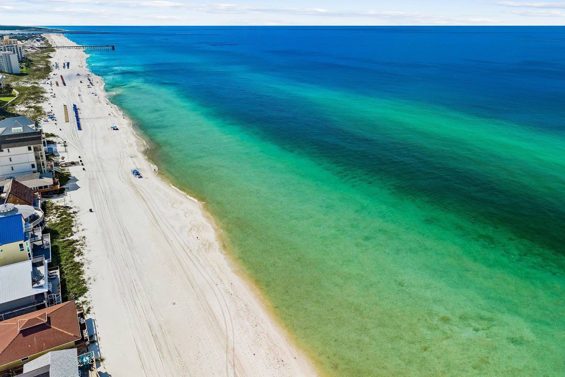 An aerial view of a beach with clear water, a pier, and buildings along the shore.
