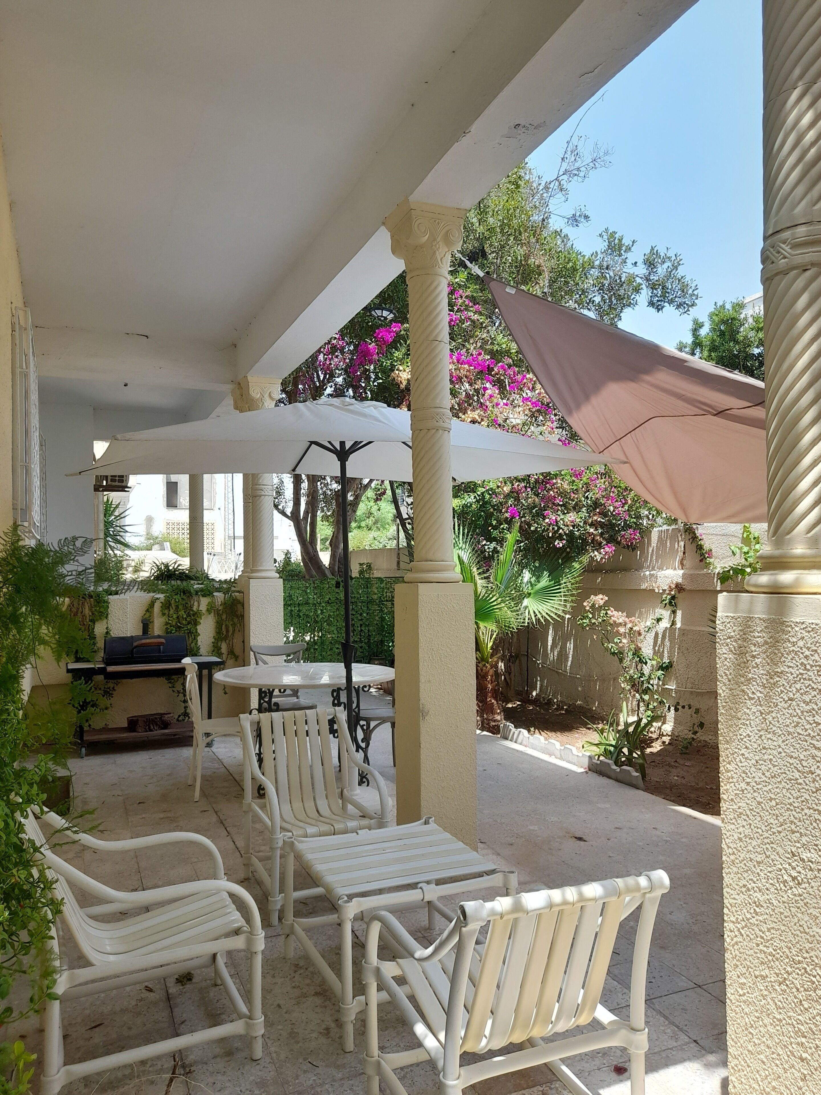 Un espace extérieur aménagé en terrasse, avec des chaises et des tables en métal blanc, un barbecue et une pergola surmontée d’une toile de couleur marron.
