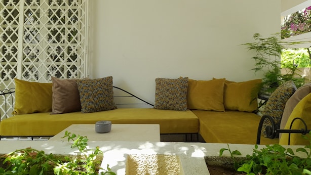 A yellow sofa with patterned cushions, a white table, and a potted plant in a sunlit room with a white lattice partition. - The House of Olive Trees (La Marsa)