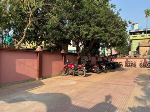 Un estacionamiento pavimentado con motocicletas y bicicletas, rodeado de árboles y un edificio de fachada colorida.