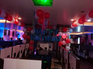 Una habitación con una mesa central rodeada de sillas, decorada con globos rojos y blancos y un cartel que dice 'HAPPY BIRTHDAY'.