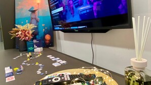 Un televisor de pantalla plana montado en la pared mostrando un programa, una mesa con varios controles remotos, una bandeja con fichas de juego y un jarrón decorativo.