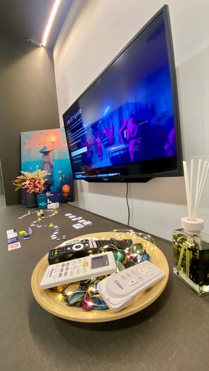 Ein flachbildfernseher an der Wand, der eine Sendung zeigt, ein Tisch mit verschiedenen Fernbedienungen, eine Ablage mit Spielsteinen und eine dekorative Vase.