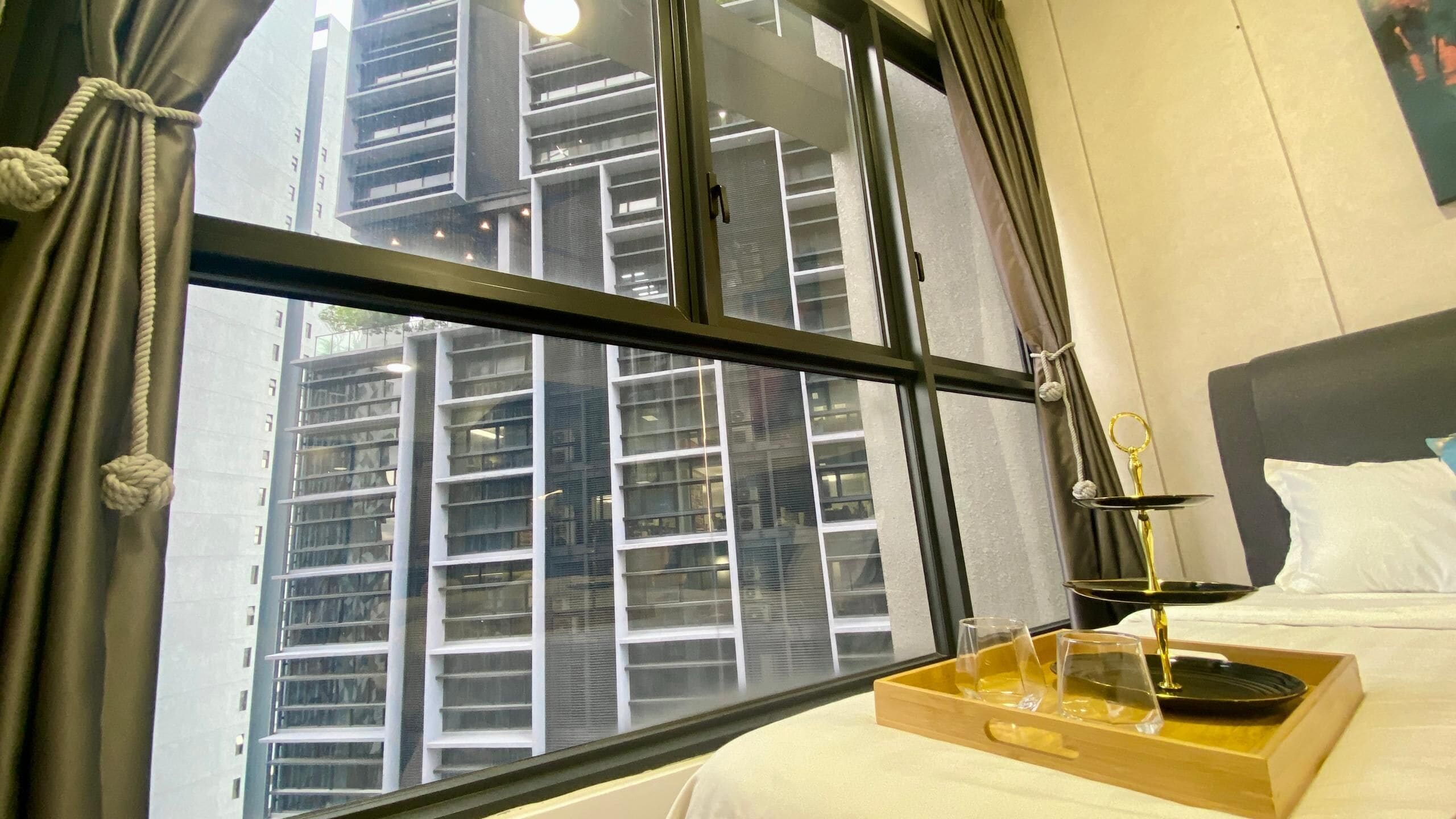 Habitació d'hotel amb una finestra gran, un llit amb roba de llit blanca, una bandeja amb vasos i una ampolla, i vista d'un edifici alt.