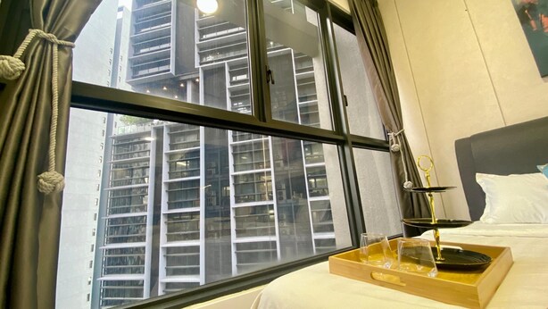 Habitación de hotel con un ventanal grande, una cama con ropa blanca, una bandeja con vasos y una botella, y vistas de un edificio alto.