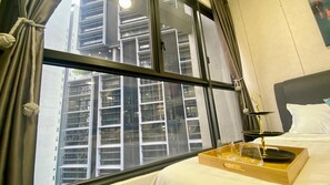 Une chambre d’hôtel avec une grande fenêtre, un lit avec une literie blanche, un plateau avec des verres et une bouteille, et une vue sur un grand immeuble.