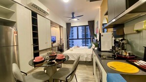 Habitación de hotel compacta con cama, comedor con mesa para dos, kitchenette y vista panorámica de la ciudad a través del ventanal.