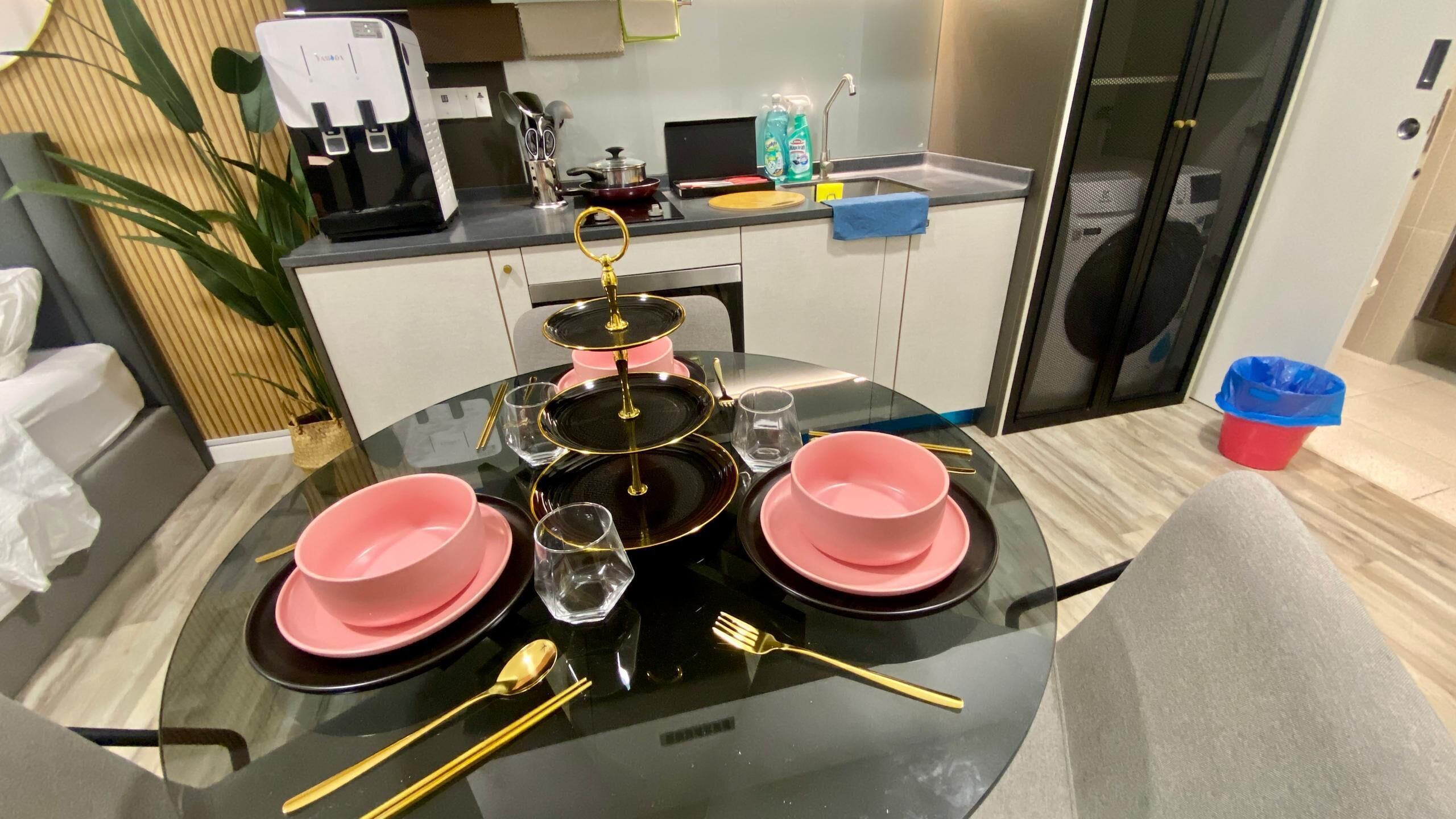 Comedor con mesa de vidrio redonda dispuesta para cuatro personas, con platos rosas y cubiertos dorados.
