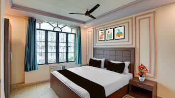 Quarto de hotel com cama, mesa de cabeceira, janela com cortinas, ventilador de teto e obras de arte nas paredes.