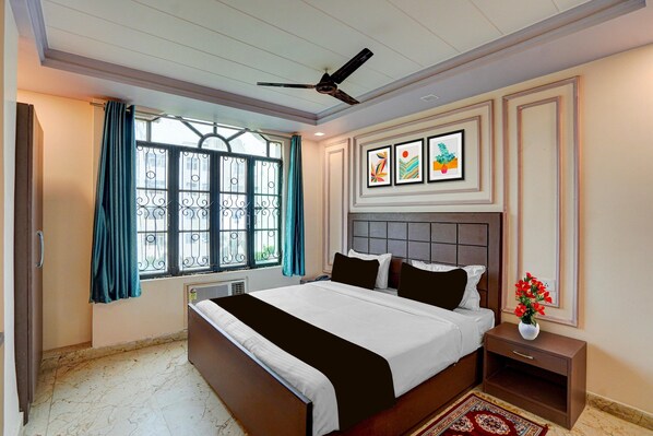 A hotel room with a bed, a nightstand, a window with curtains, a ceiling fan, and wall art. - Super Deluxe Room Mauji's (Prayagraj)
