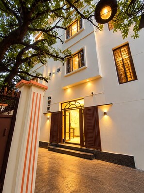 Un edificio de dos pisos con una entrada bien iluminada, una escalera que conduce a la puerta y un árbol en primer plano.