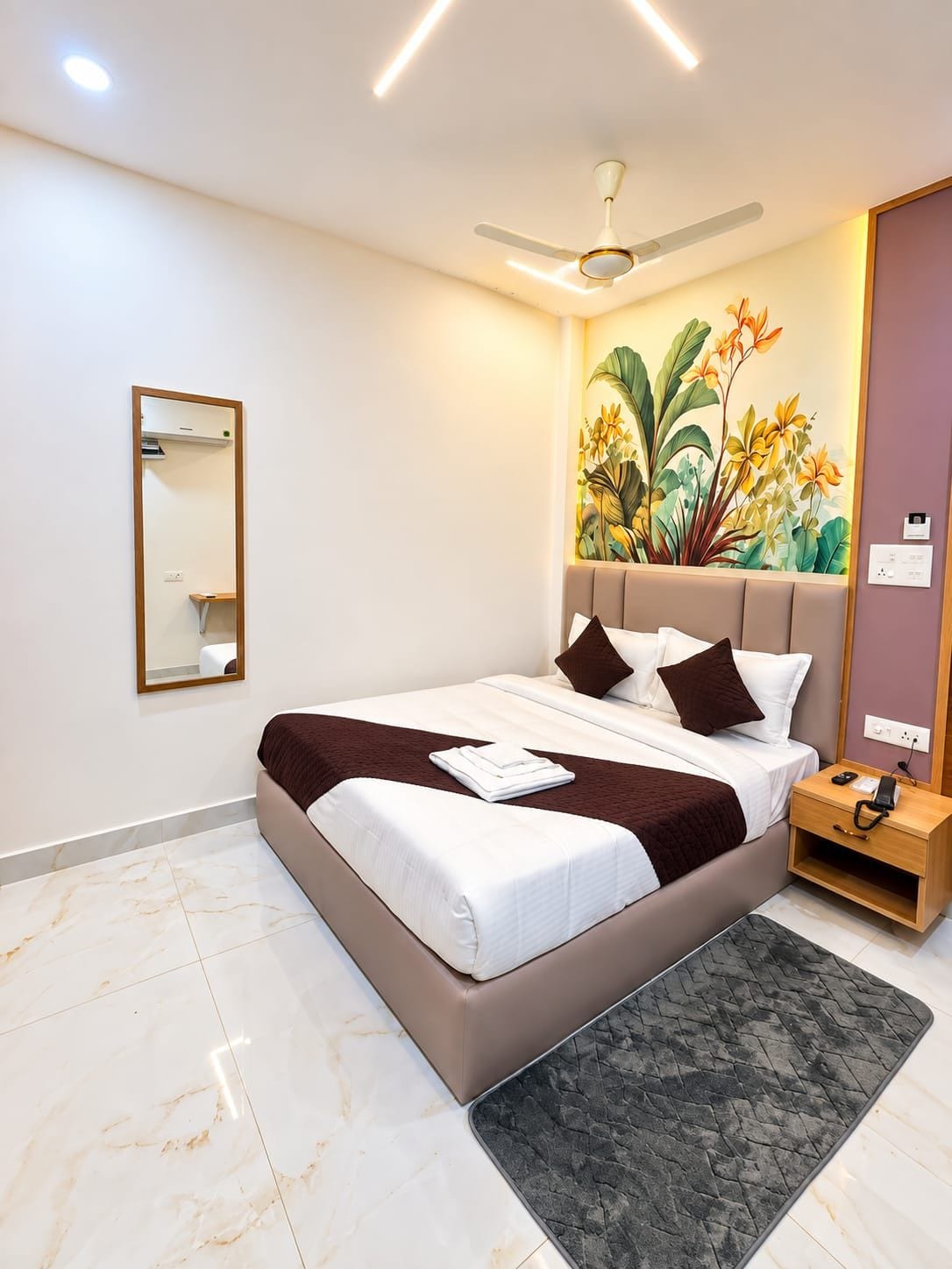 Una habitación de hotel con una cama, una mesita de noche, un ventilador de techo, un espejo montado en la pared y un adorno floral en la pared.