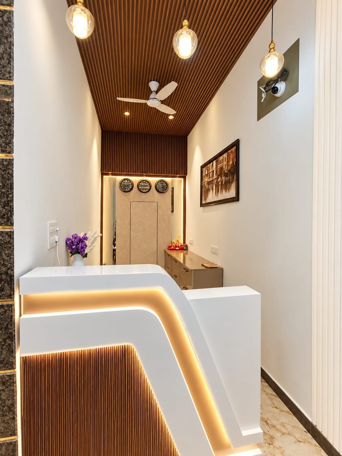 Un espace d’accueil au design moderne, comprenant un comptoir de réception, un ventilateur de plafond, des horloges murales et un tableau encadré.