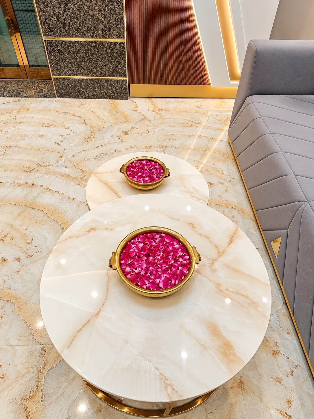 A marble-topped table with two gold-rimmed bowls filled with pink flower petals, set on a marble floor with a grey upholstered sofa in the background.