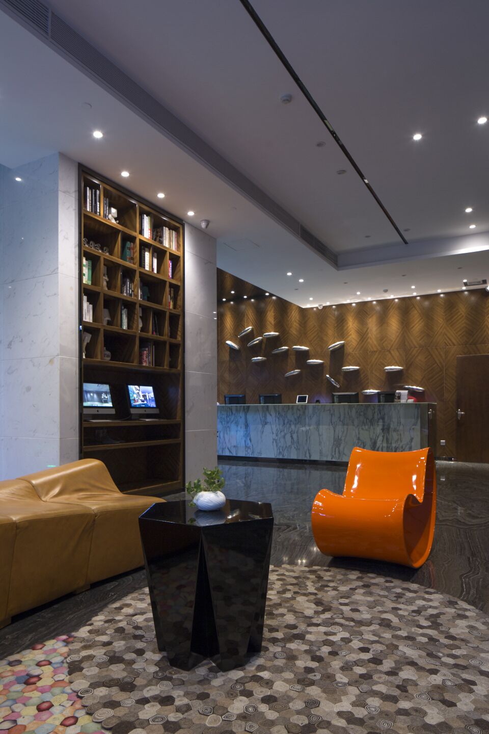Un hall con mostrador de recepción, una estantería, un sofá de cuero, un sillón naranja y un suelo estampado.
