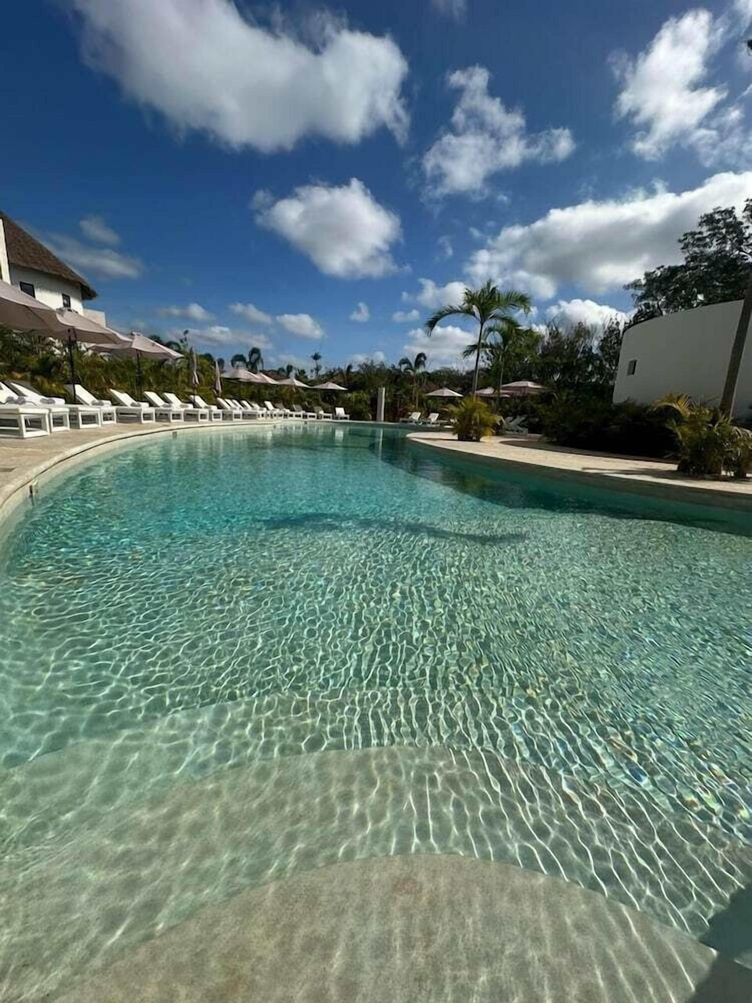 Una piscina cristalina con sillas de descanso y sombrillas, rodeada de edificios y vegetación.