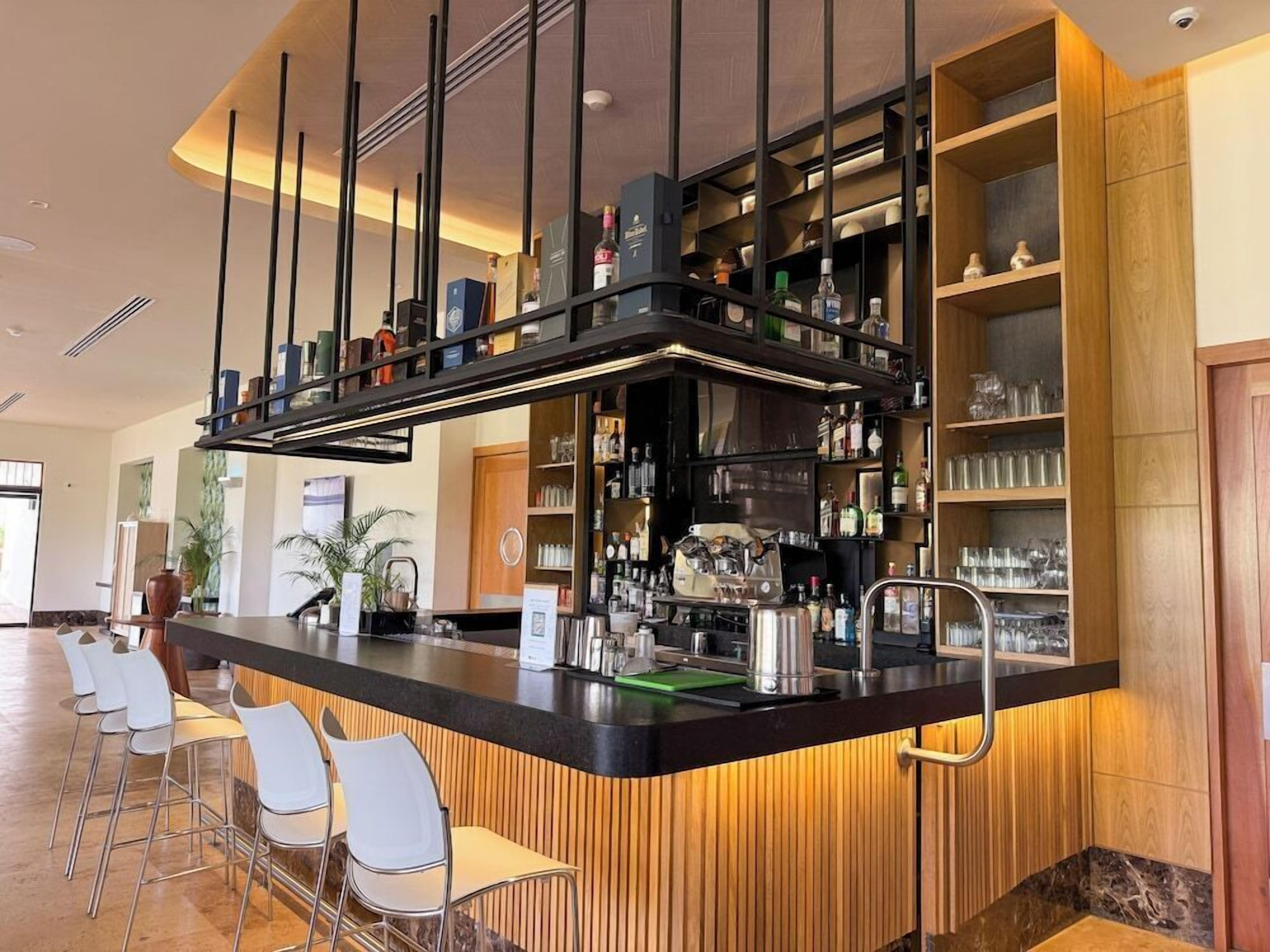 Una zona de bar con mostrador, taburetes y estantes llenos de botellas y vasos.