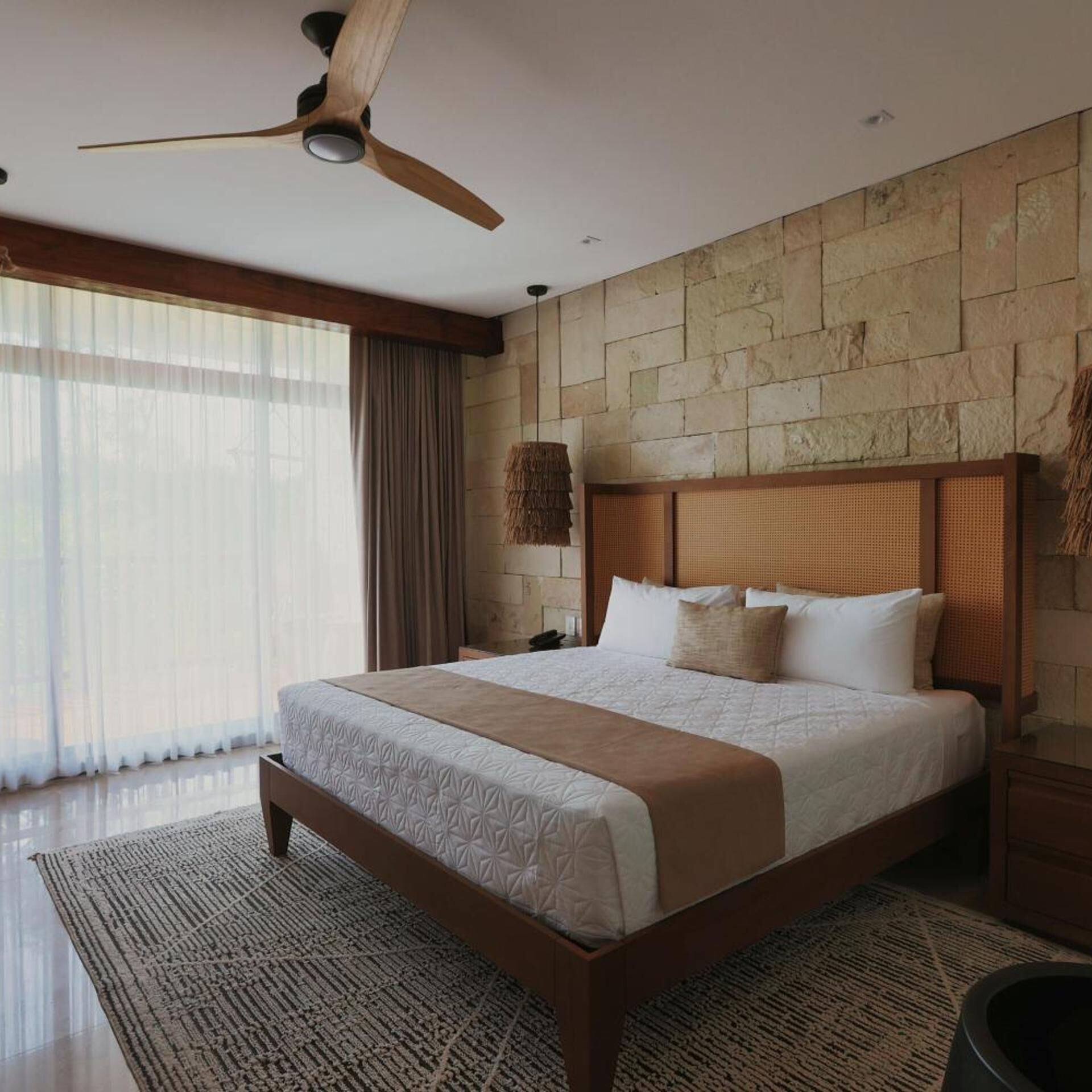 Un dormitorio amplio con una cama grande, ventilador de techo, pared de piedra y vistas al exterior.