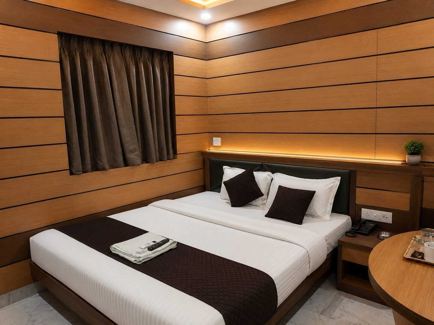 Ein Hotelzimmer mit einer Wandpaneele aus Holz, einem Bett mit weißer Bettwäsche und dunklen Kissen, einem Nachttisch mit Telefon und einer kleinen Pflanze sowie einem runden Tisch mit Tablett.