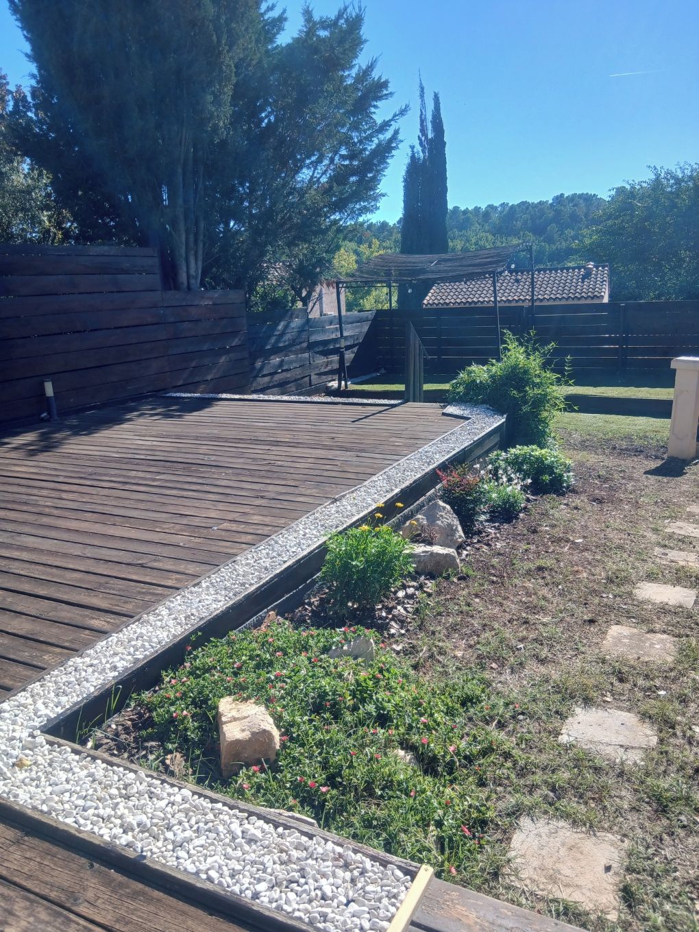 A wooden deck with a gravel border, surrounded by a grassy area with plants and rocks.