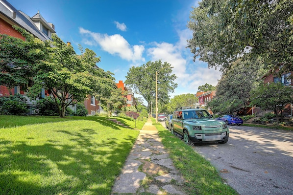 A residential street with a green SUV parked on the side, a sidewalk, and houses with brick facades.