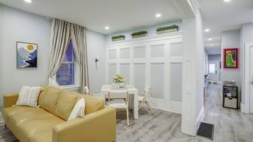 Un salon moderne avec un canapé jaune, une table à manger avec des chaises, un meuble blanc et un couloir menant à une autre pièce.