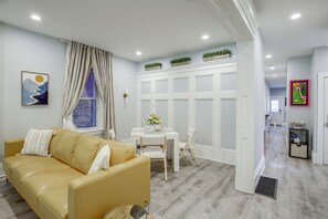 A modern living room with a yellow sofa, a dining table with chairs, a white cabinet, and a hallway leading to another room.