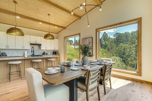 A modern kitchen with wooden cabinets, a large island with bar stools, and a dining area with a view of the forest. - Brand-New Smokies Cabin w/ Spectacular Mtn Views (Sevierville)