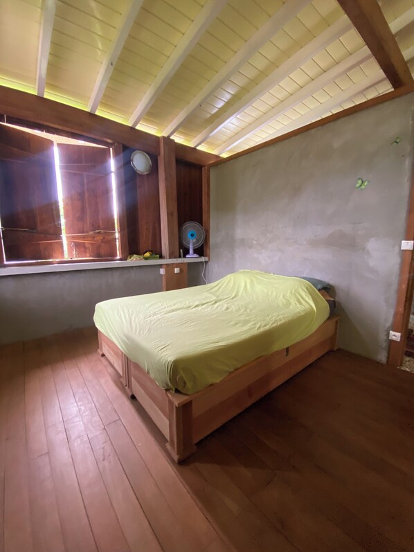 A bedroom with a wooden bed, a window with blinds, a wall-mounted fan, and a ceiling with wooden beams.