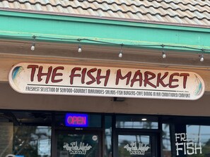 Ein Geschäft mit einer Schaufensterfront, auf der der Schriftzug "The Fish Market" zu sehen ist. Es werden Meeresfrüchte, Marinaden, Salate, Fischburger und Cafébetrieb in klimatisierter Umgebung angeboten.