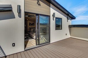 A modern building with a sliding glass door leading to a furnished interior, a wooden deck, and a clear sky. - Game Room & Stunning Mtn Views! Cedar City Home (Cedar City)