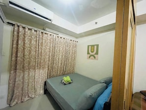 A bedroom with a bed, curtains, an air conditioning unit, and a wall-mounted artwork. - Charming 1-Bedroom Apartment Near Mall of Asia (Pasay)