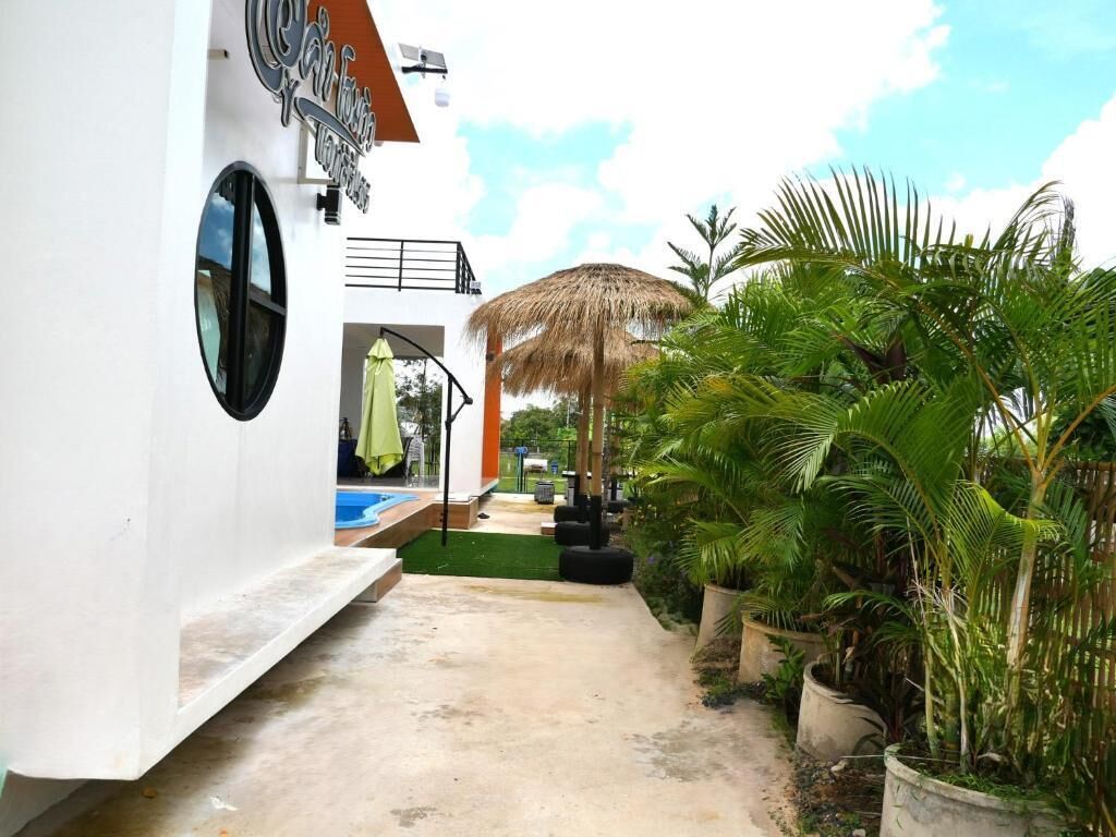 A white building with a circular window and a sign. There is a concrete pathway leading to a pool area with a thatched umbrella.