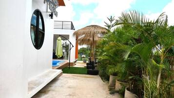 A white building with a circular window and a sign. There is a concrete pathway leading to a pool area with a thatched umbrella.