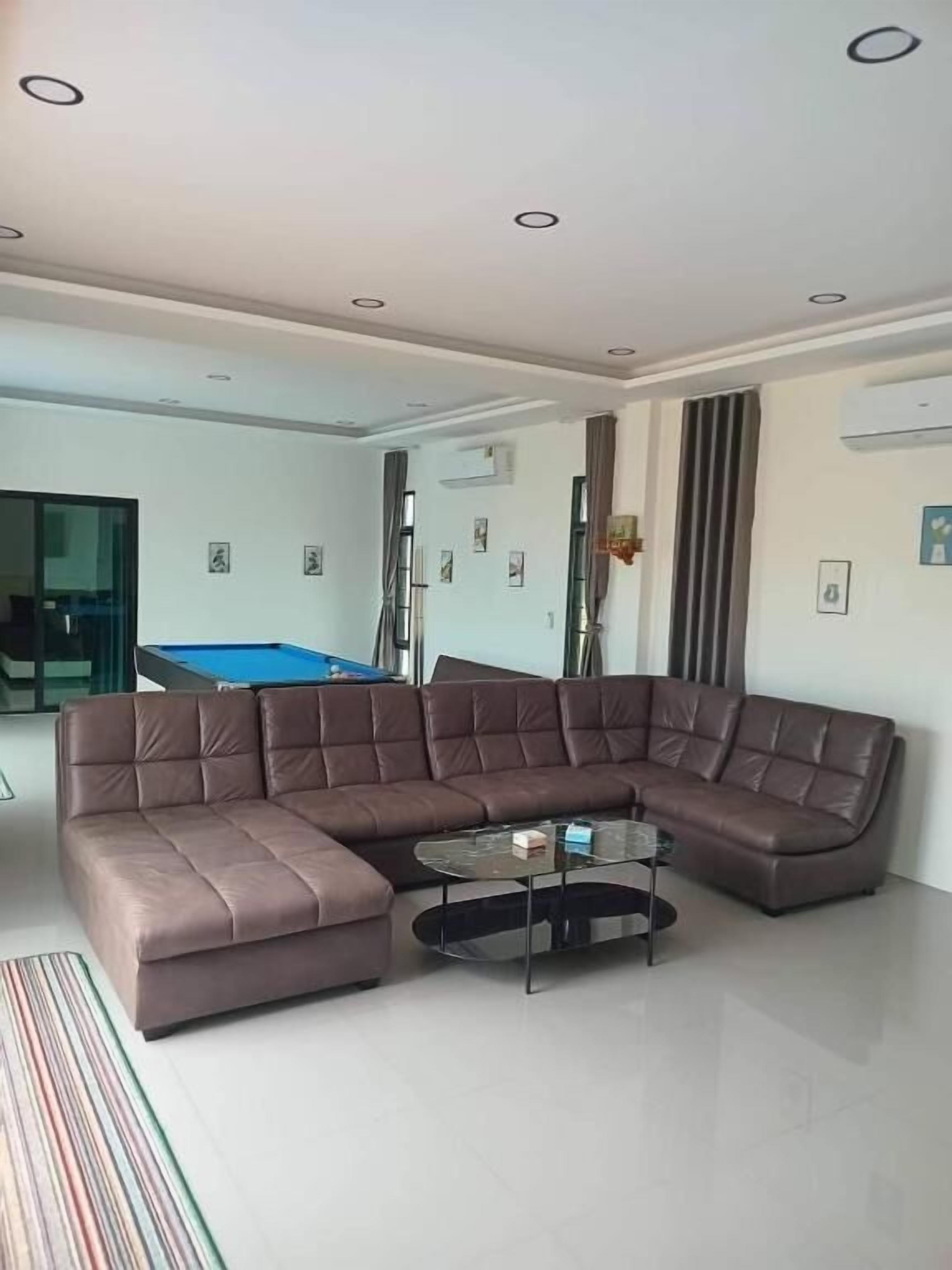 A modern living room with a sectional sofa, a glass coffee table, and a pool table in the background.