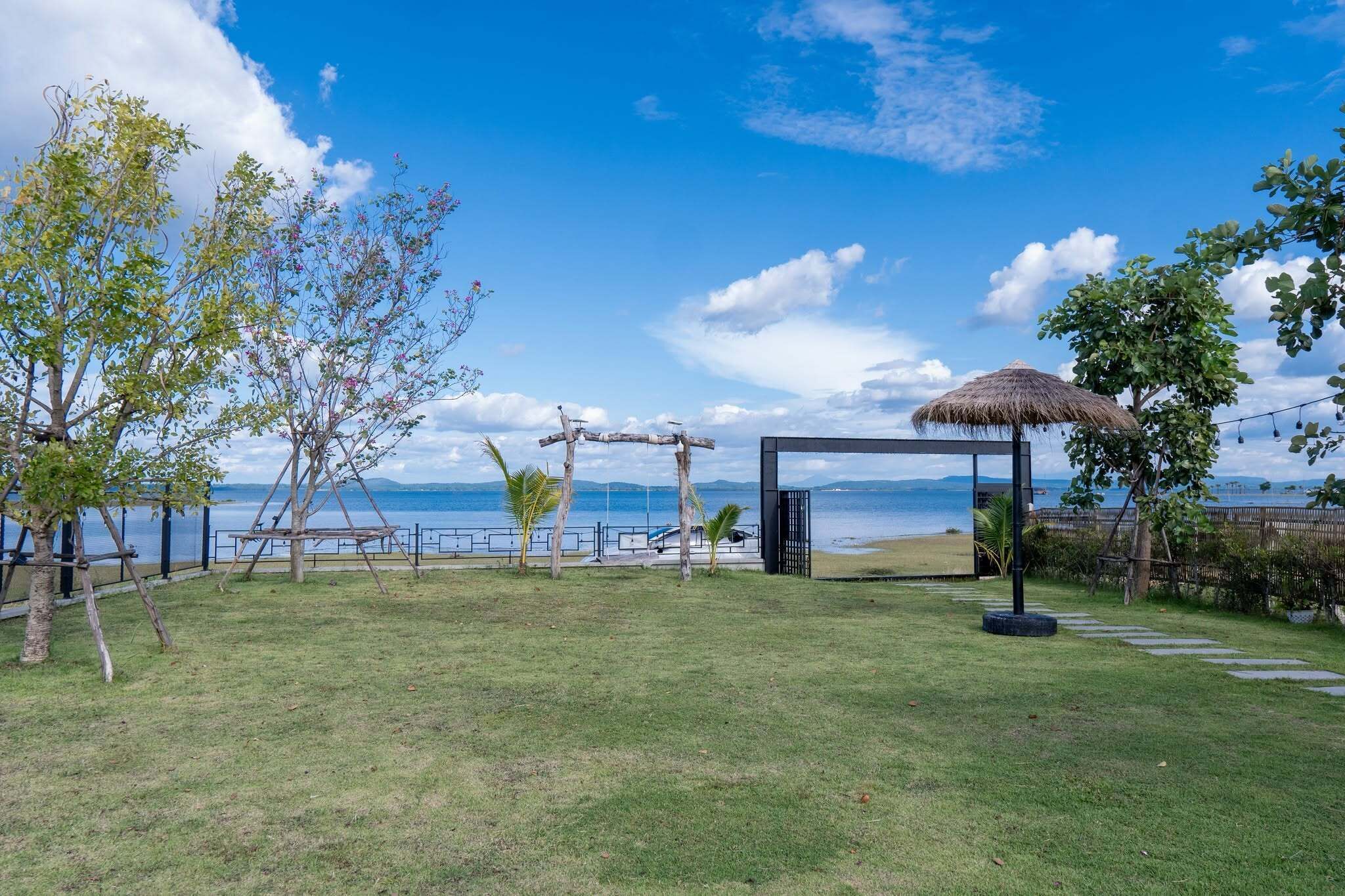 Un espacio al aire libre con césped, un columpio de madera, una estructura de techo de paja y vista al mar.