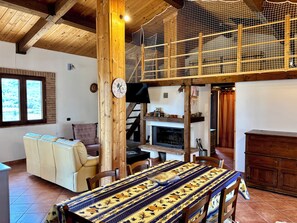 A cozy living room with a fireplace, a dining table with a patterned tablecloth, and a wooden ceiling.