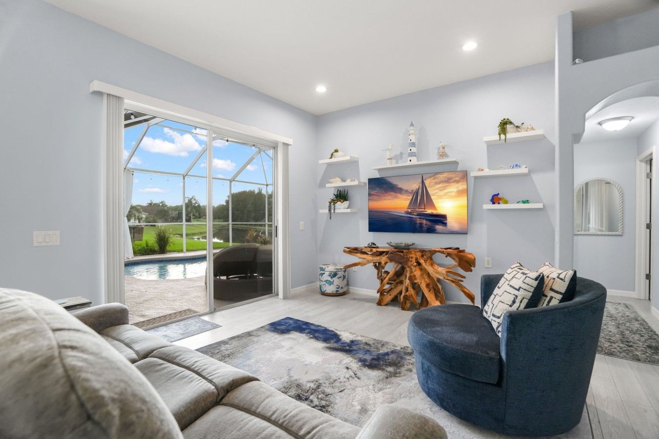 A modern living room with a large glass door leading to a pool, a blue armchair, a wooden coffee table, and a painting of a sailboat.