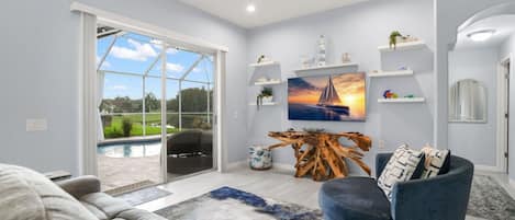 A modern living room with a large glass door leading to a pool, a blue armchair, a wooden coffee table, and a painting of a sailboat.