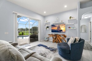 Un salon moderne doté d’une grande porte vitrée donnant sur une piscine, d’un fauteuil bleu, d’une table basse en bois et d’un tableau représentant un voilier.
