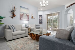 A modern living room with a glass coffee table, two sofas, and a view of the outdoors through a sliding glass door.