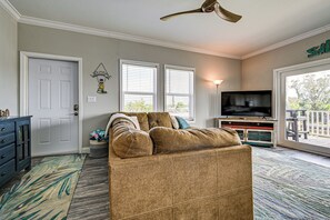 Ein Wohnzimmer mit braunem Sofa, Deckenventilator, Fernseher auf einem Holzboden und Blick ins Freie durch eine Schiebetür.