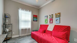 Ein Zimmer mit rotem Sofa, einem Fenster mit Jalousien, einem Regal mit Dekoration und Wandbildern.