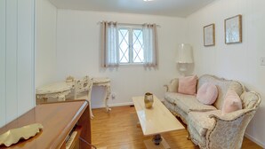 Et værelse med trægulv, et hvidt skrivebord med stol, en sofa med puder, et vindue med gardiner og en loftlampe.
