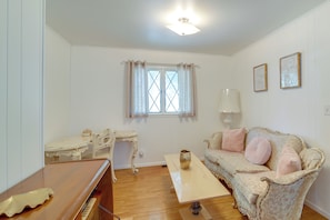 A room with a wooden floor, a white desk with a chair, a sofa with cushions, a window with curtains, and a ceiling light. - Backyard Oasis! Spacious Tulsa Home w/ Pool (Tulsa)