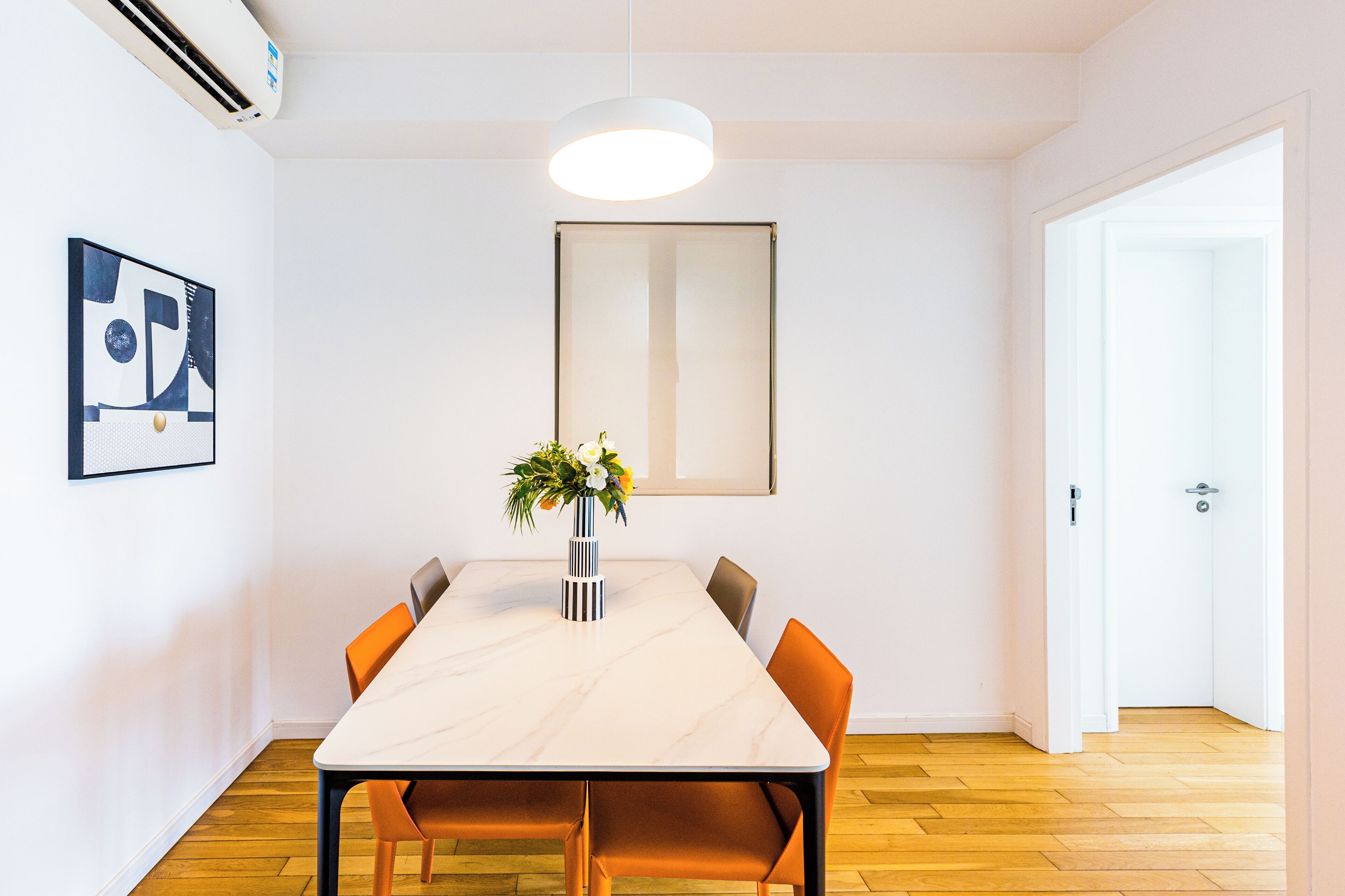 A dining area with a marble table, orange chairs, a vase with flowers, a pendant light, and a framed abstract artwork on the wall.