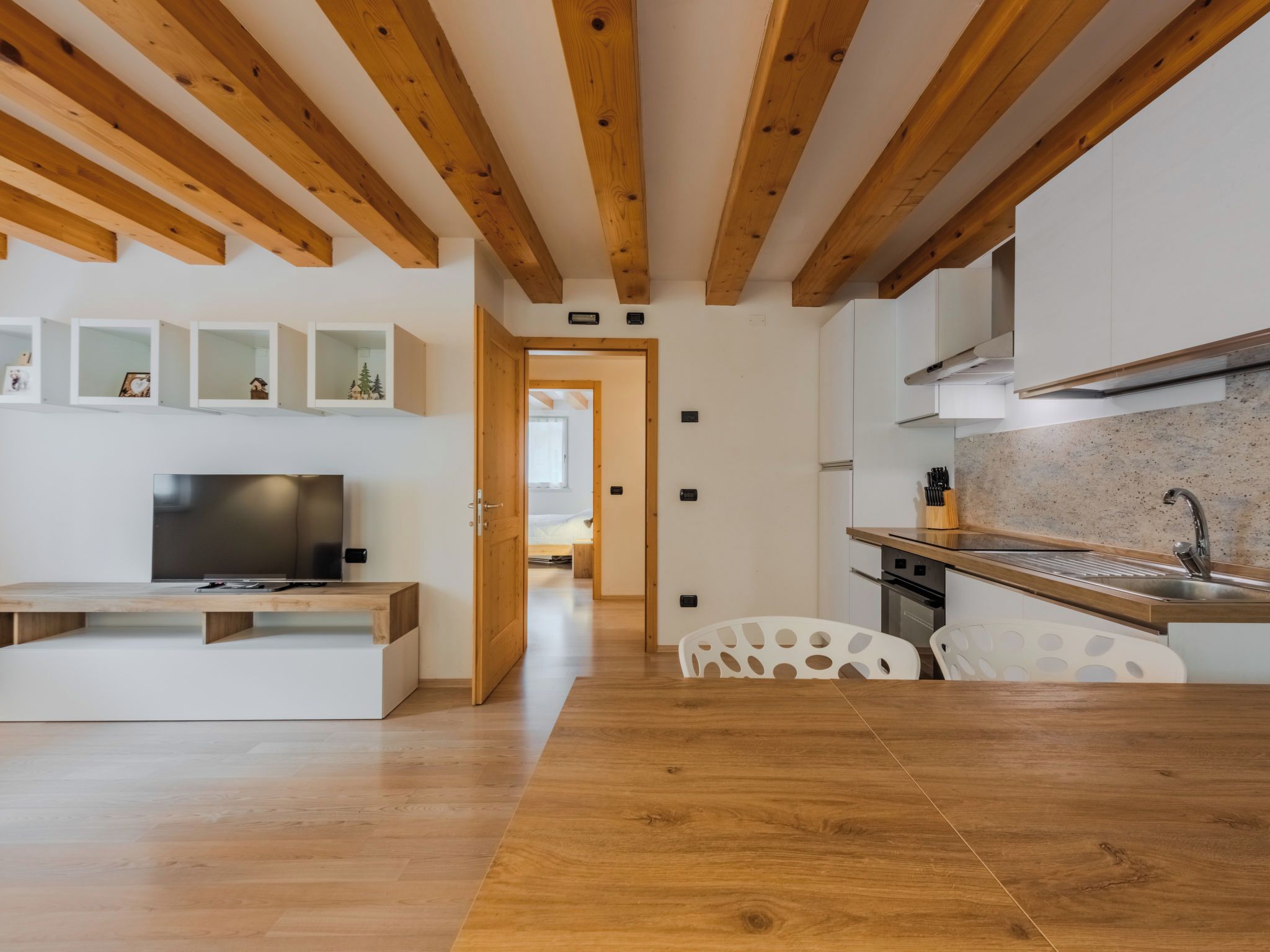 Una cocina moderna con vigas de madera, armarios blancos y una zona de comedor con mesa y sillas de madera.